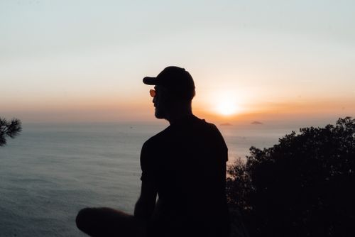 Free Back view silhouette of relaxed male in cap and sunglasses sitting on tranquil seacoast and admiring scenic sunset over rippling calm seawater Stock Photo