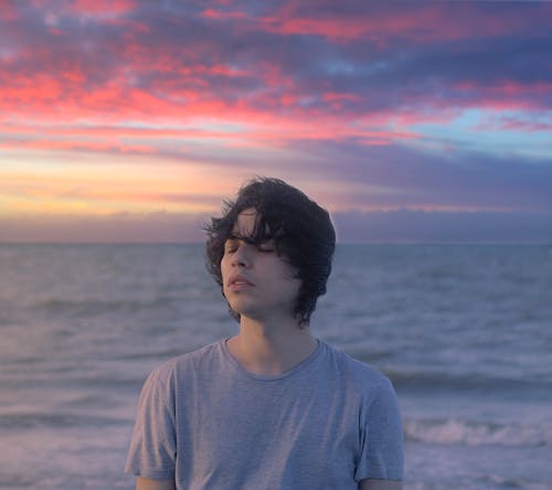 Calm young male with closed eyes in casual clothes enjoying fresh breeze on coast of sea