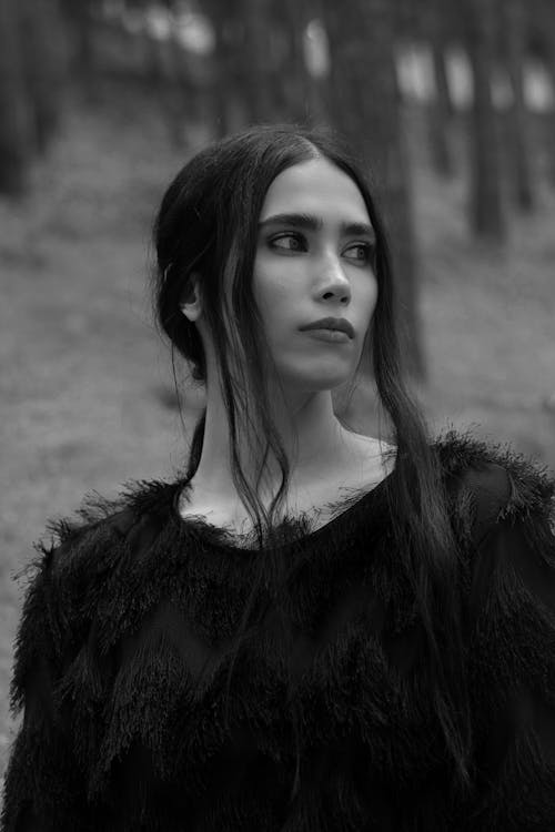 Black and white attractive calm female with long dark hair wearing fluffy sweater standing in woods and looking away