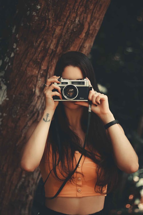 Unrecognizable female photographer wearing top looking looking through vintage photo camera and taking shot while standing tree trunk in nature