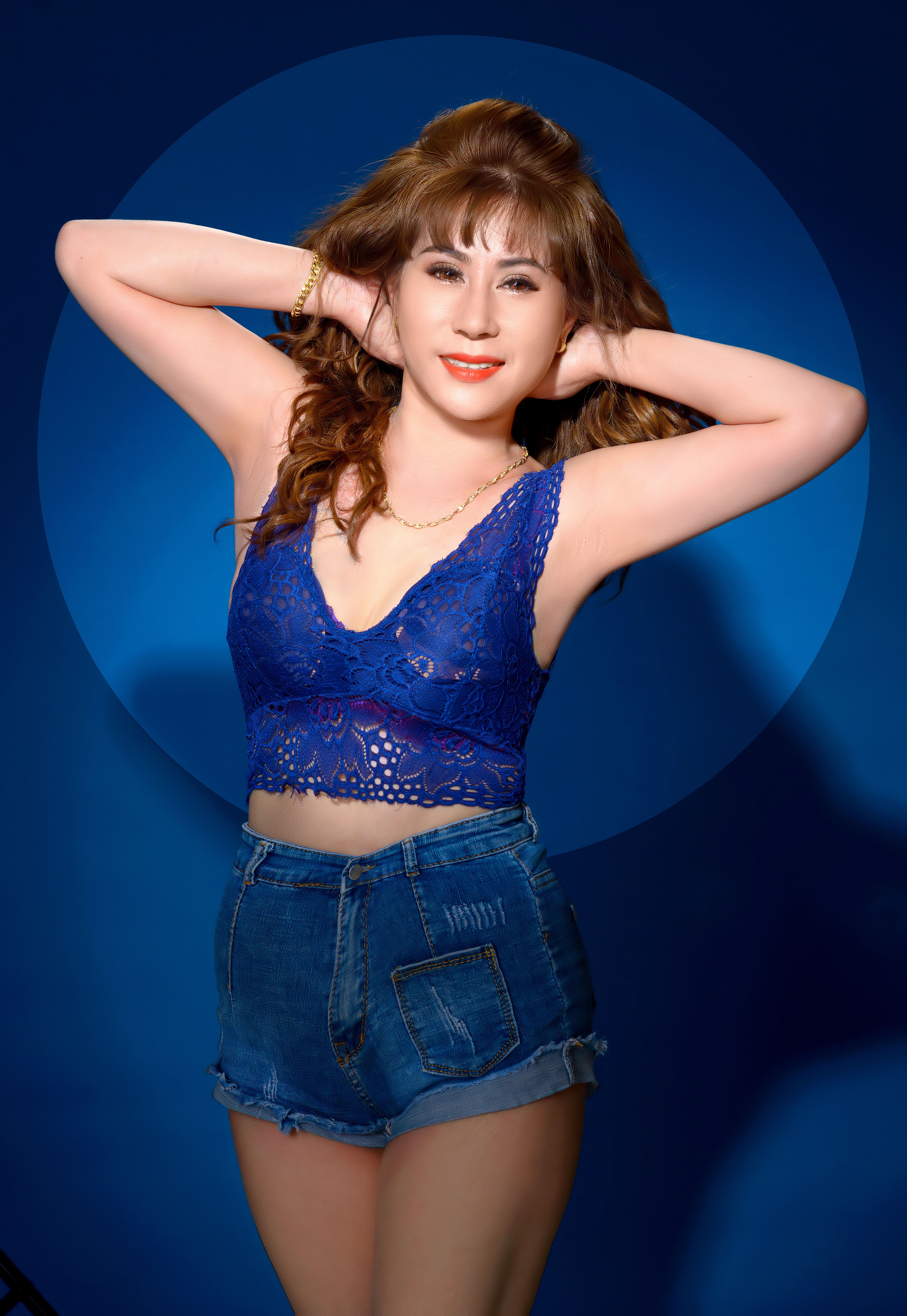 Smiling woman in a blue lace top and denim shorts posing in a studio.