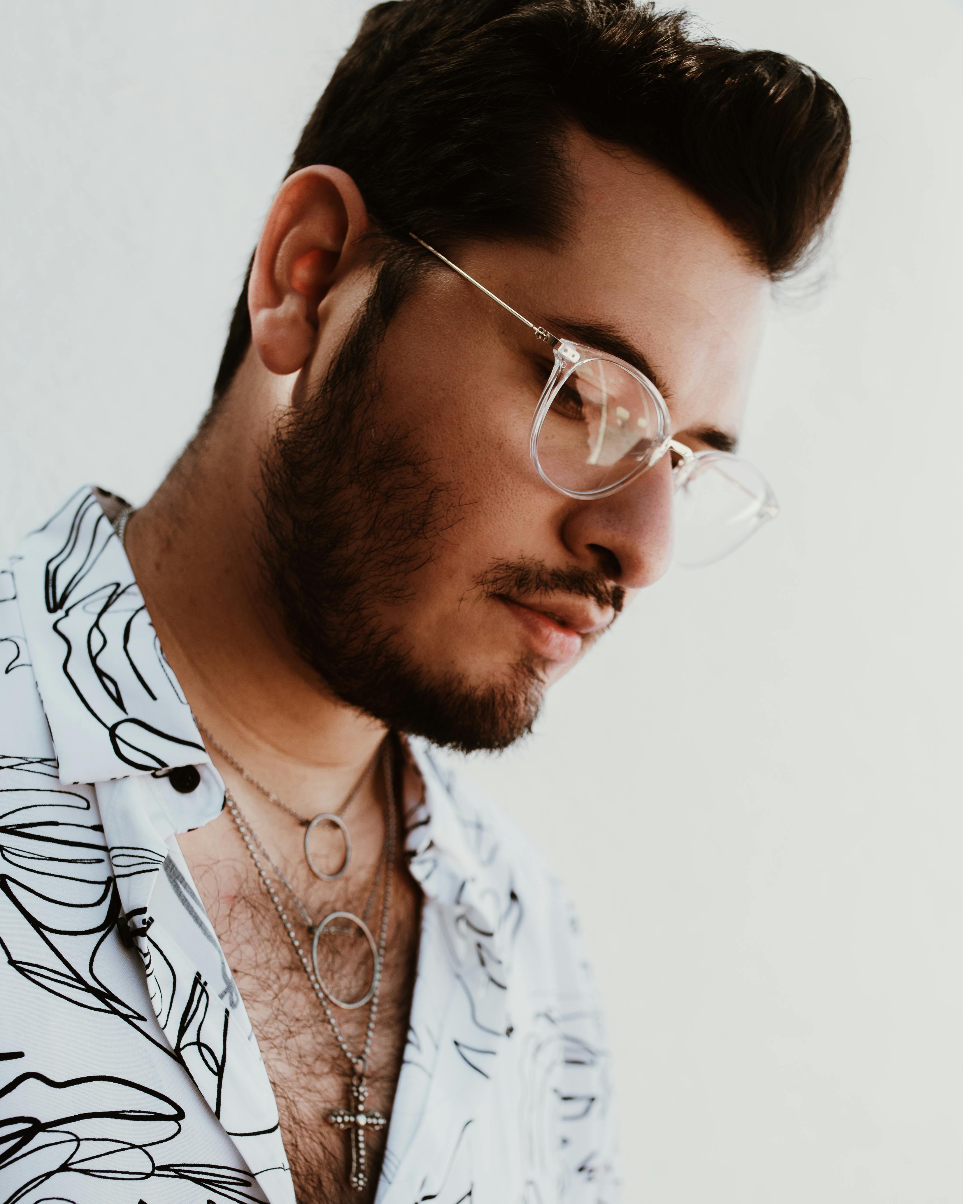 Portrait of a fashionable man in glasses wearing a patterned shirt and necklaces, looking down.