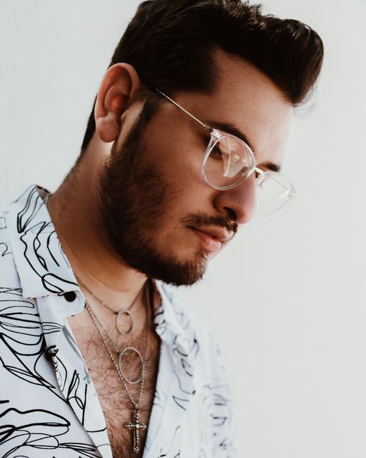 Portrait of a fashionable man in glasses wearing a patterned shirt and necklaces, looking down.