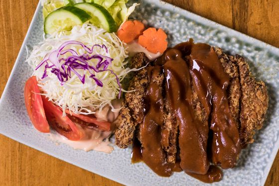 Breaded pork cutlet covered in brown sauce beside shredded cabbage, tomato and cucumber