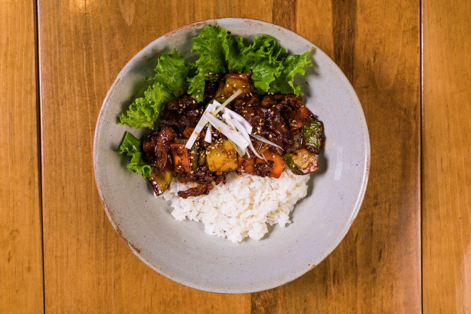 A glossy reddish-brown Korean BBQ sauce in a small white bowl, studded with sesame seeds, with a spoon lifting a thick ribbon of sauce and grilled ribs in the background.