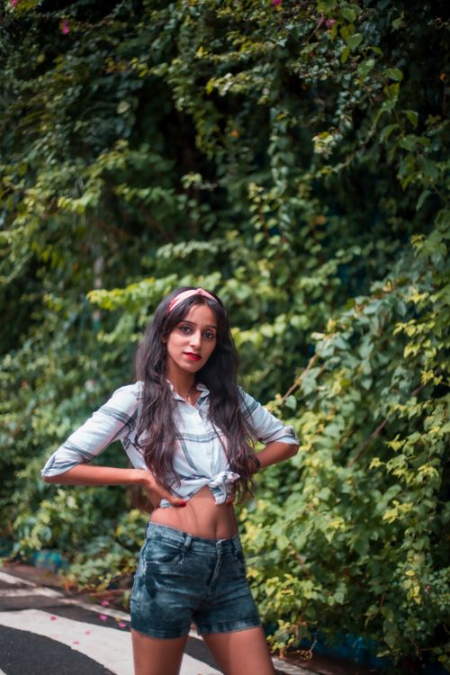 Free Stylish woman in denim shorts and plaid shirt posing outdoors with lush green plant background. Stock Photo