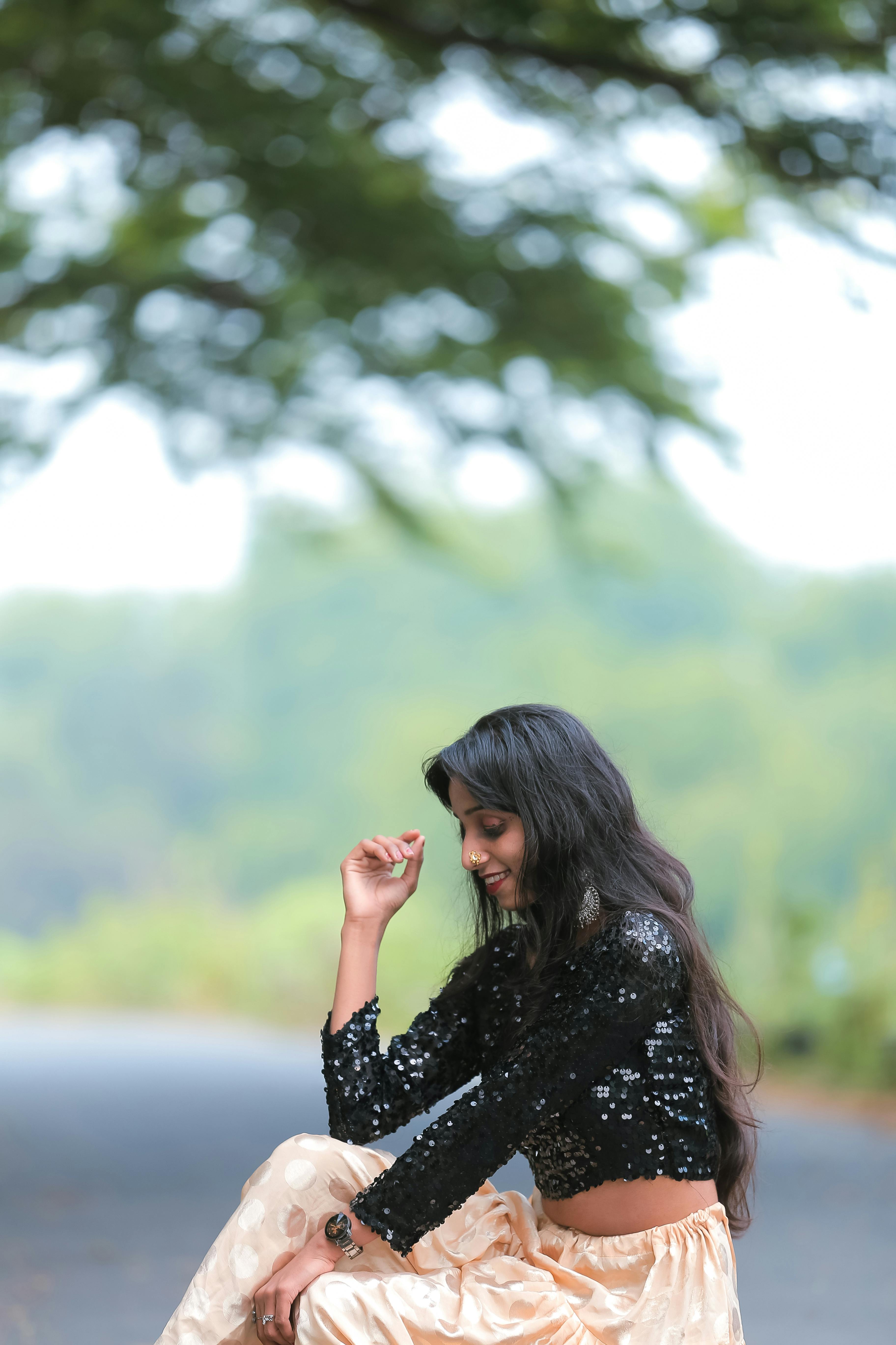 Woman sitting outdoors on a road in vibrant greenery, wearing a black sequin top.