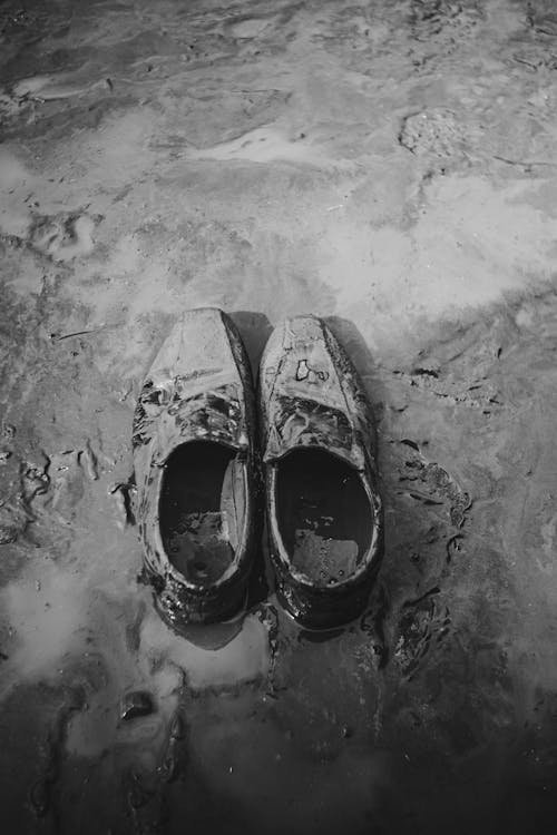 From above black and white of aged male footwear with mud on roadway with clay