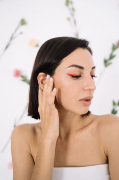 Close-up of a woman with striking red eye makeup, touching her face against a floral backdrop.
