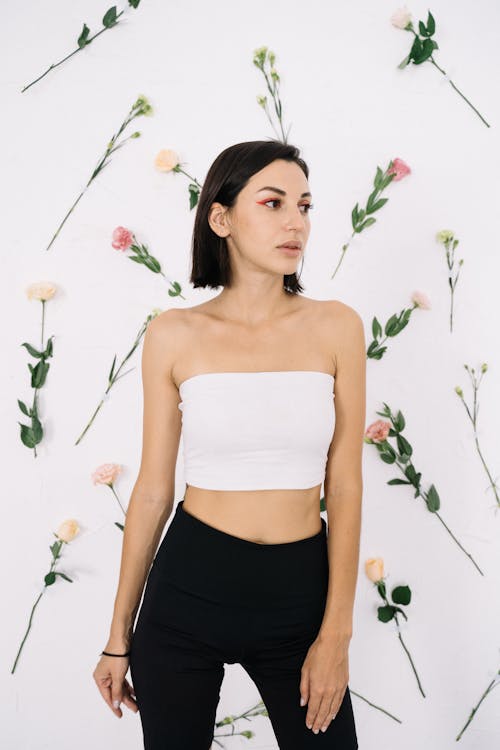 Free Fashionable woman in black pants and tube top posing against white wall decorated with flowers. Stock Photo
