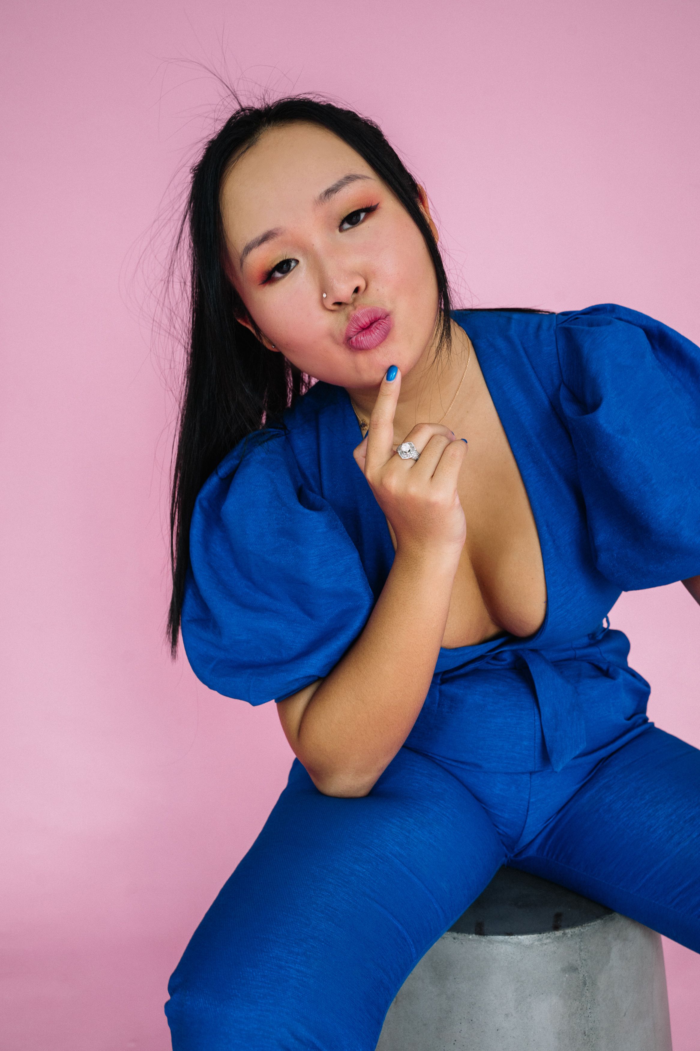 Asian woman striking a playful pose in a blue outfit against a pink background.