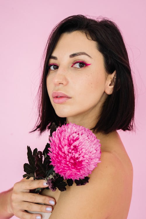 A Woman in Short Hair Holding a Stem of Pink Aster Flower while Looking at the Camera