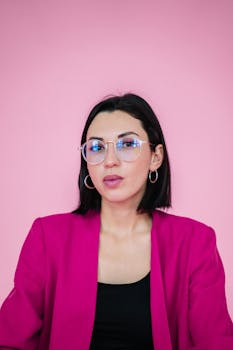 Portrait of a woman in a pink blazer with eyeglasses, set against a pink background.