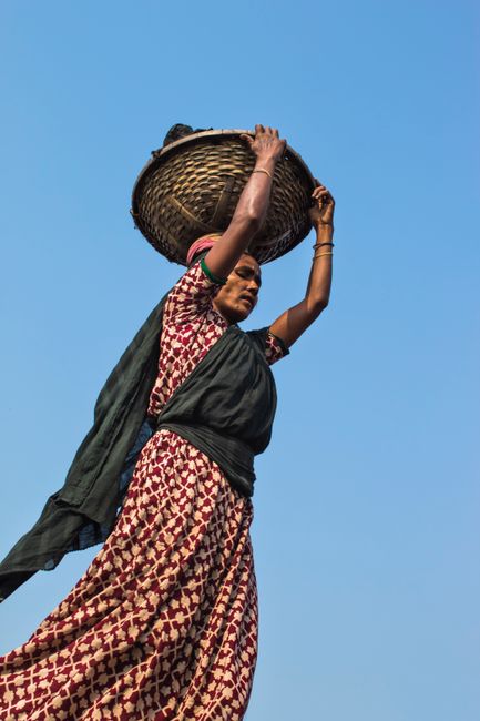 A woman in patterned dress lifts a basket overhead against a clear blue sky.