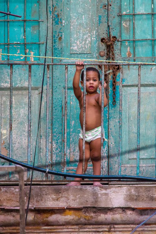Free A child standing behind a metal railing in front of a weathered turquoise door. Stock Photo