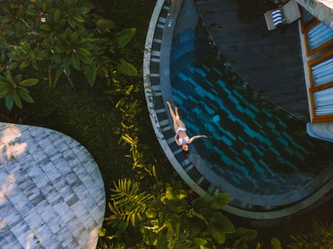Aerial view of a woman in a bikini relaxing by a swimming pool surrounded by lush greenery in Bali.