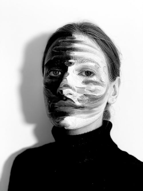 Black and white of young serious female model covering face with contrast paints looking at camera in light studio