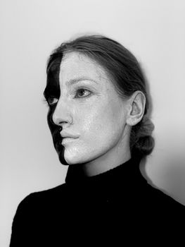 Black and white of thoughtful weird female in turtleneck with painted face looking up while standing on white background in room