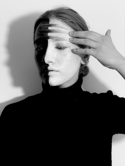 Black and white of strange mysterious female wearing turtleneck smearing paints on face with hand while looking down on white background
