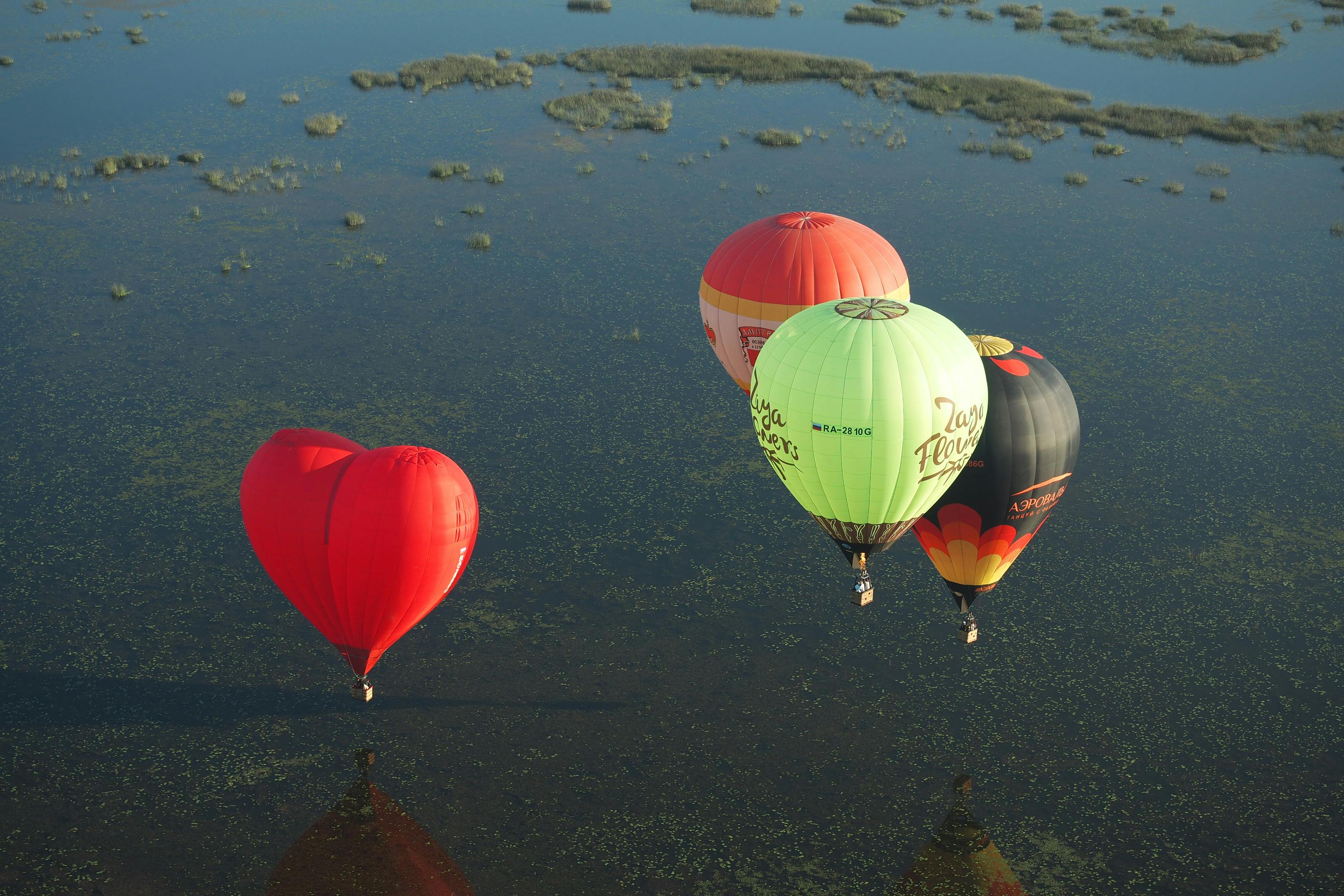 Four colorful hot air balloons, including a heart-shaped one, float over a calm lake.