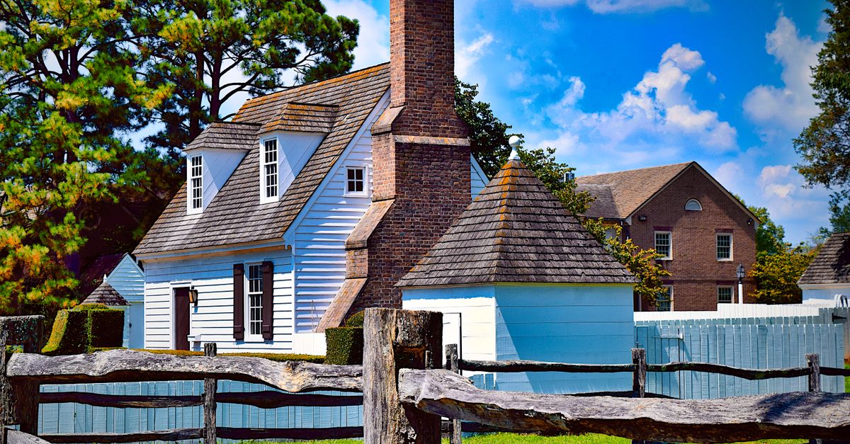 Beautiful colonial-style house with a wooden fence and lush greenery under a bright blue sky.