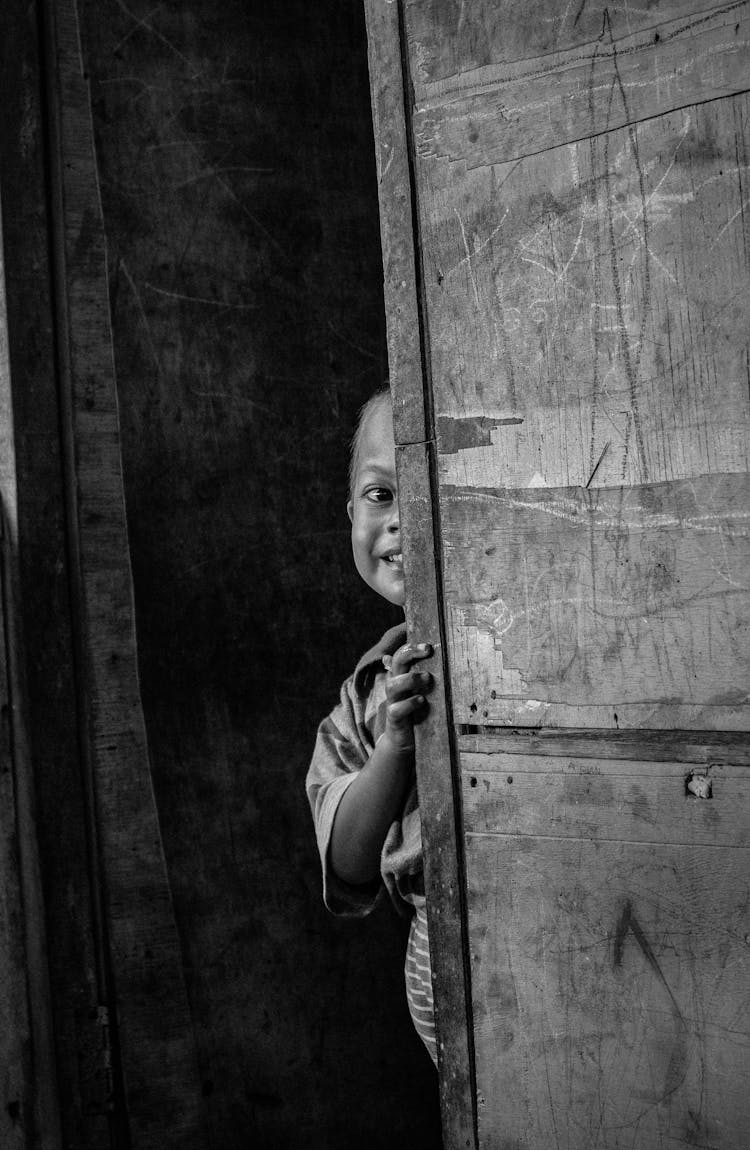 A Grayscale Photo Of Child Hiding On A Wooden Door