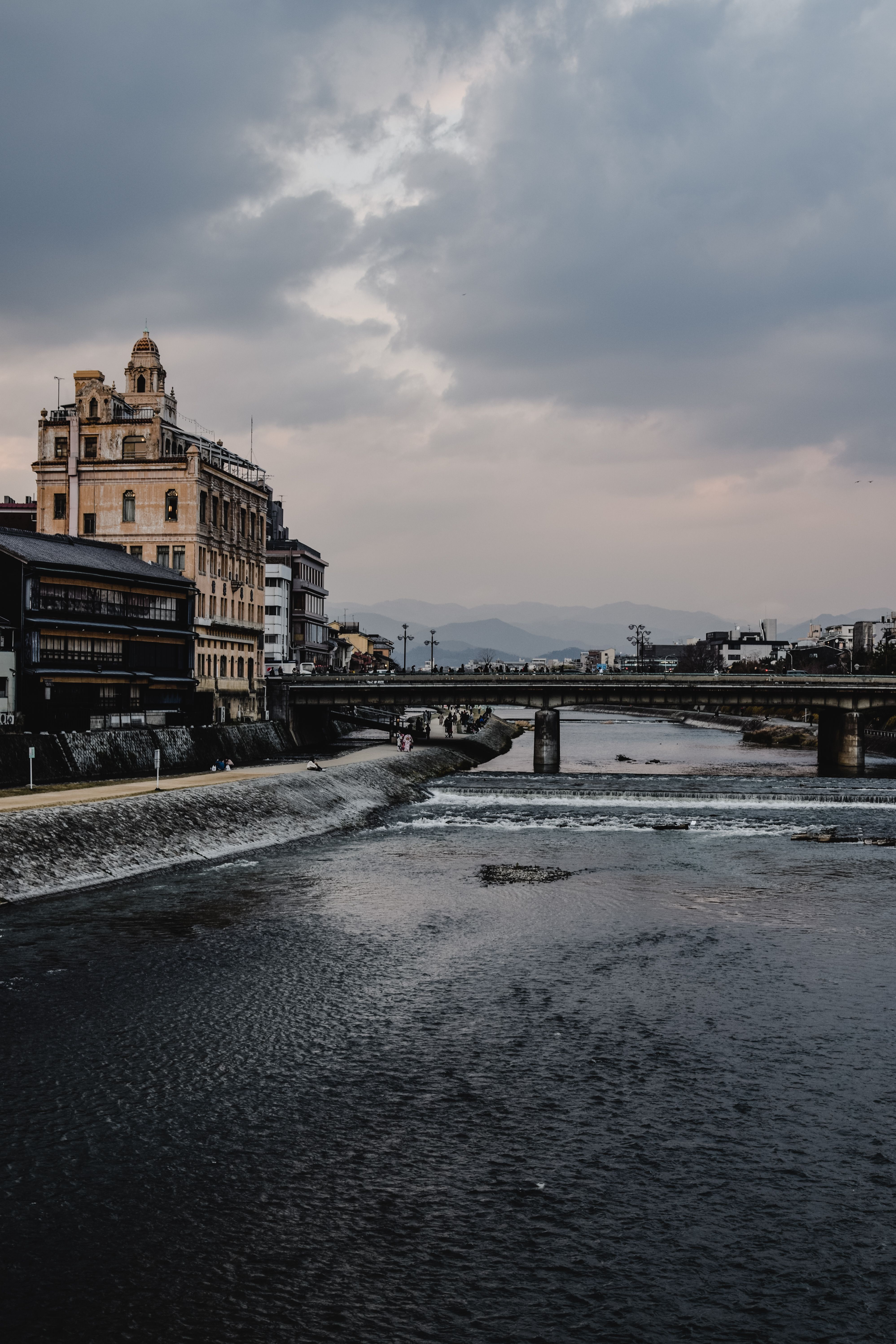 京都河畔的建筑在阴云密布的天空下，与宁静流淌的河水共同构成了一幅富有戏剧性的景色