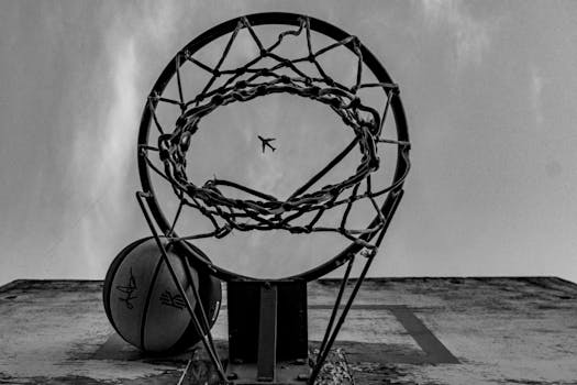 Black and white image capturing a unique view of a basketball hoop with an airplane in the sky.