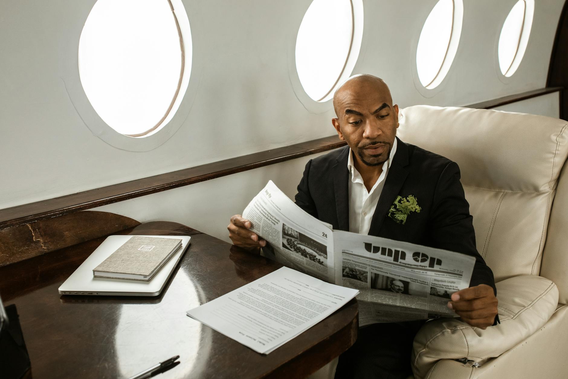 African American man reading newspaper in luxury private jet, wearing a black suit and white shirt.