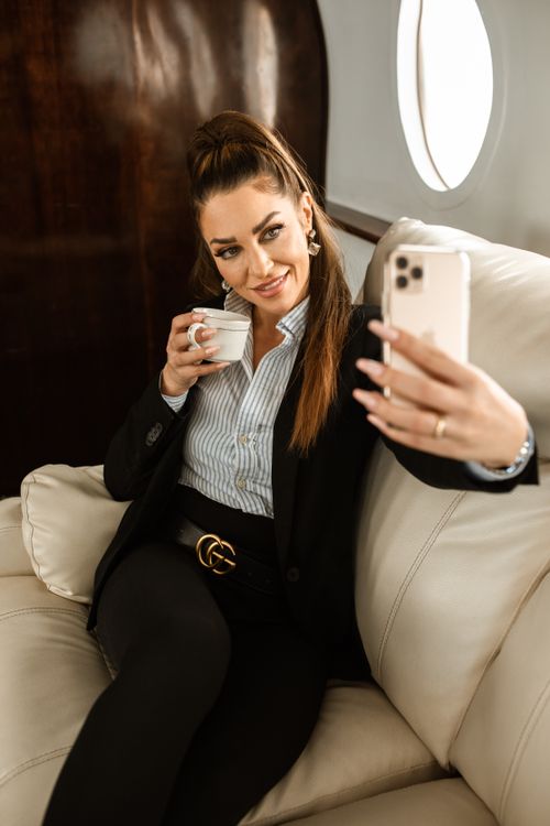 Free Stylish businesswoman relaxes with coffee, capturing a selfie on a private jet. Stock Photo