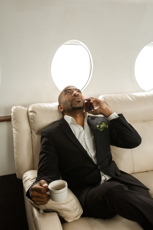 Free Businessman in a black suit talks on his phone while relaxing in a private jet with a cup of coffee. Stock Photo