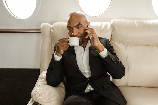 Elegant businessman seated in private jet, drinking coffee and on a phone call.