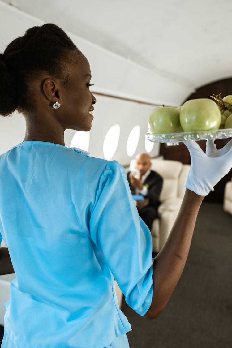 Backview Of A Flight Attendant Carrying Serving Tray Full Of Fruits 