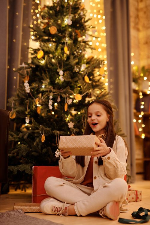 Free Happy child with present box sitting with crossed legs on floor near shiny decorative tree during New Yer holiday Stock Photo