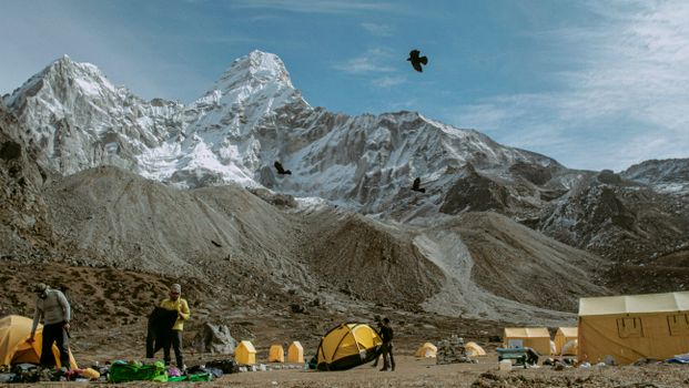 在白雪皑皑的山峰下，鸟儿飞翔，露营者正在风景优美的山地露营地准备帐篷