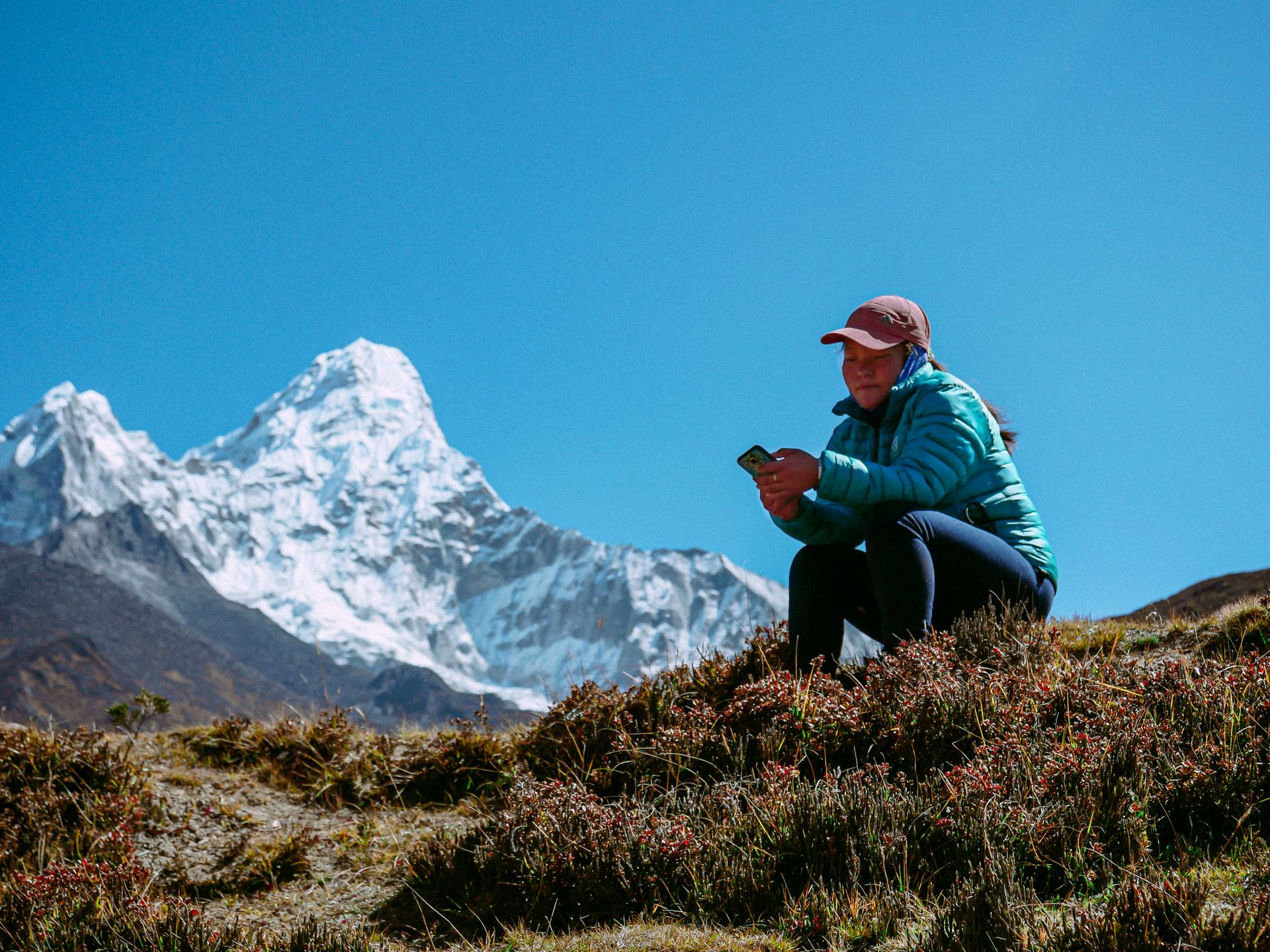 Woman sits on a hilltop using phone with Ama Dablam mountain backdrop in Nepal.
