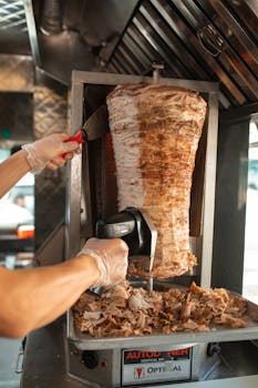 Hands slicing shawarma meat on a vertical rotisserie in a food truck.