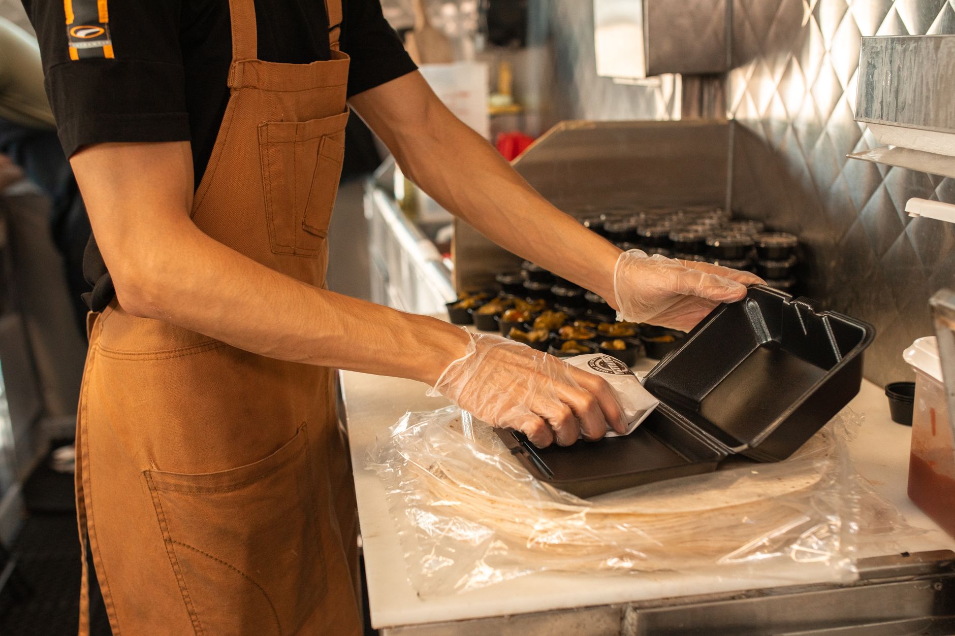 Chef wearing an apron and gloves, preparing takeaway food containers in a kitchen setting.