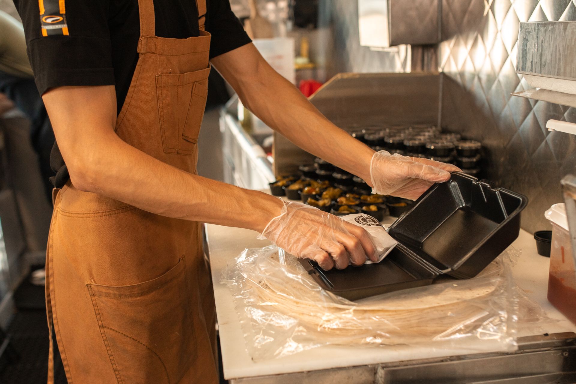 Chef wearing an apron and gloves, preparing takeaway food containers in a kitchen setting.