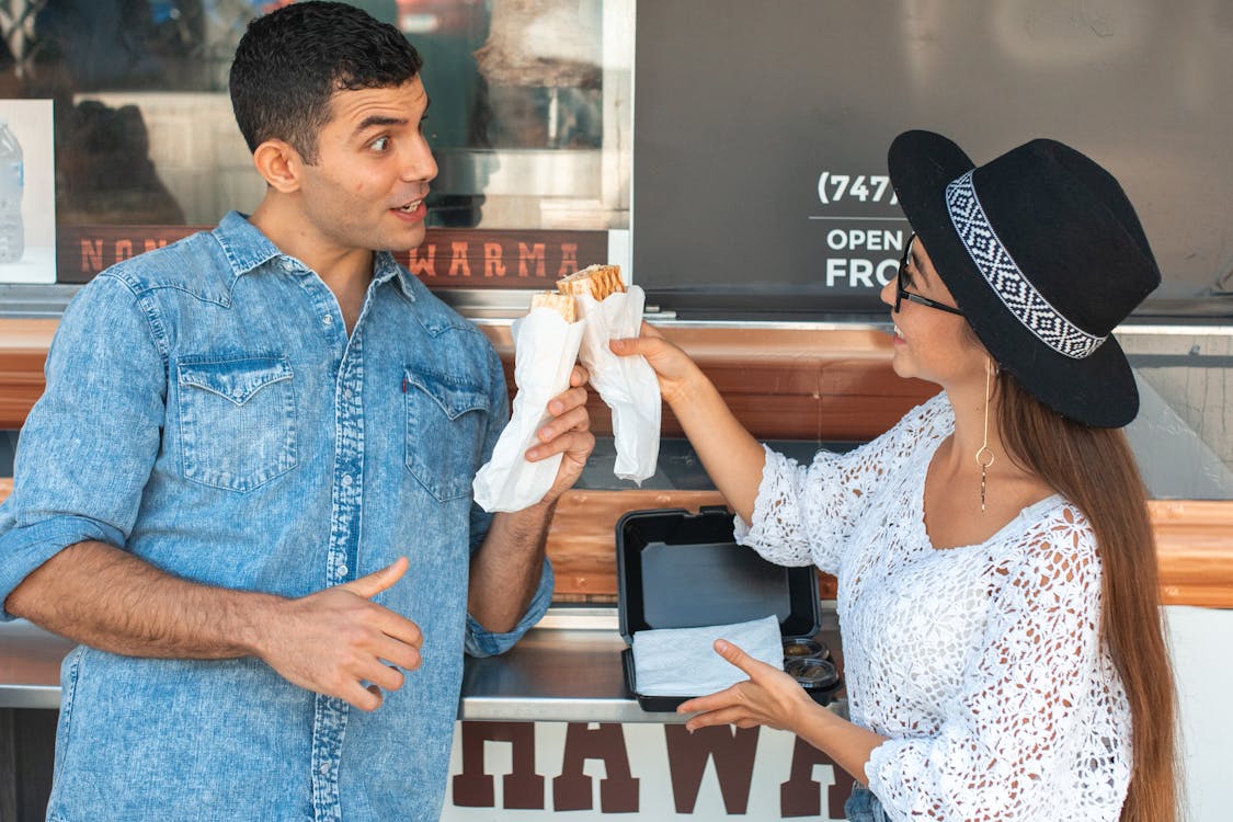 Cheerful grimacing couple in casual clothes clinking sandwiches while standing near food truck and looking at each other