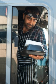Male food vendor presenting a takeout box from a food truck window, wearing plaid shirt and apron.