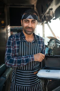 Smiling man inside a food truck wearing a striped apron, holding a food tray.