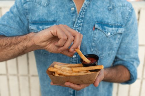 Free Close-up of a person dipping French fries in ketchup while holding a paper container. Stock Photo