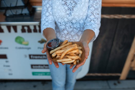 A person holds a serving of crispy French fries with ketchup outside a food stall.