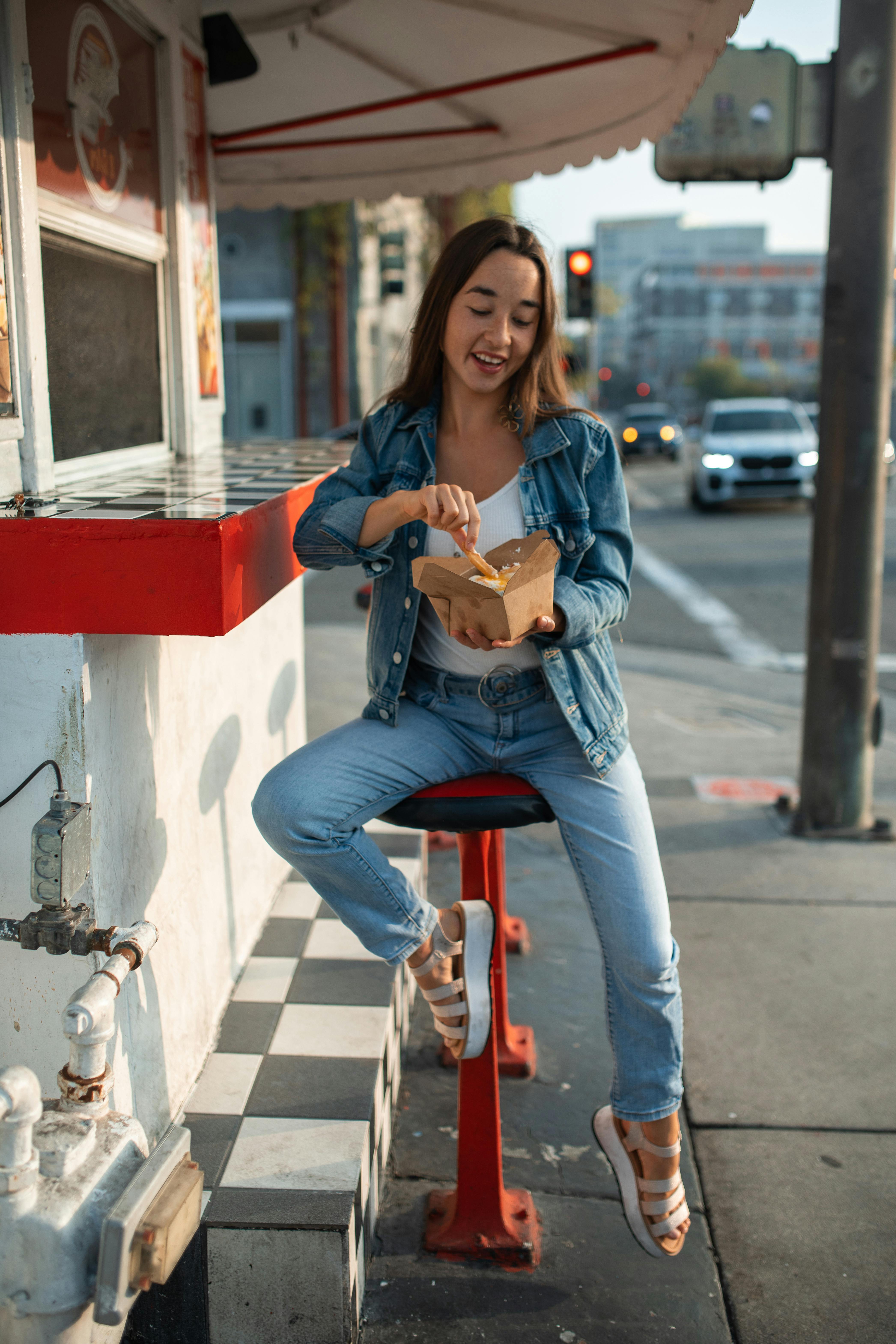 a woman in denim jeans sitting on a stool while holding a takeout box