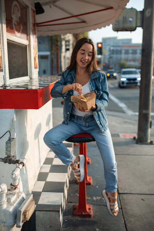 Free A Woman in Denim Jeans Sitting on a Stool while Holding a Takeout Box Stock Photo