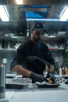 A chef in black attire prepares food inside a contemporary food truck kitchen, showcasing culinary skills.