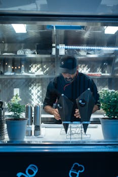 Man in food truck wearing a black cap and uniform, preparing meals with plants visible.