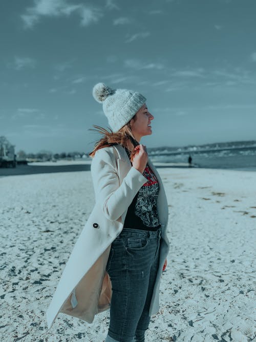 Side view of stylish female wearing warm coat and hat standing on sandy shore near water while enjoying view of sea