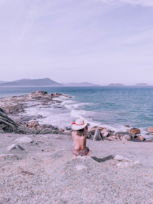 Back view of unrecognizable female with bare back wearing hat sitting on beach with pebble near waving sea and stony coast in summer
